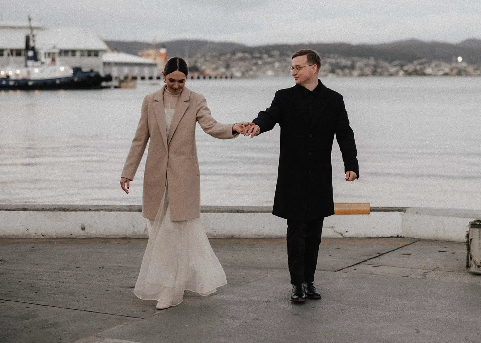 A newlywed couple in elegant wedding attire walks hand in hand along the Hobart waterfront in Tasmania, dressed warmly for the cool weather, with a scenic view of the harbour behind them.