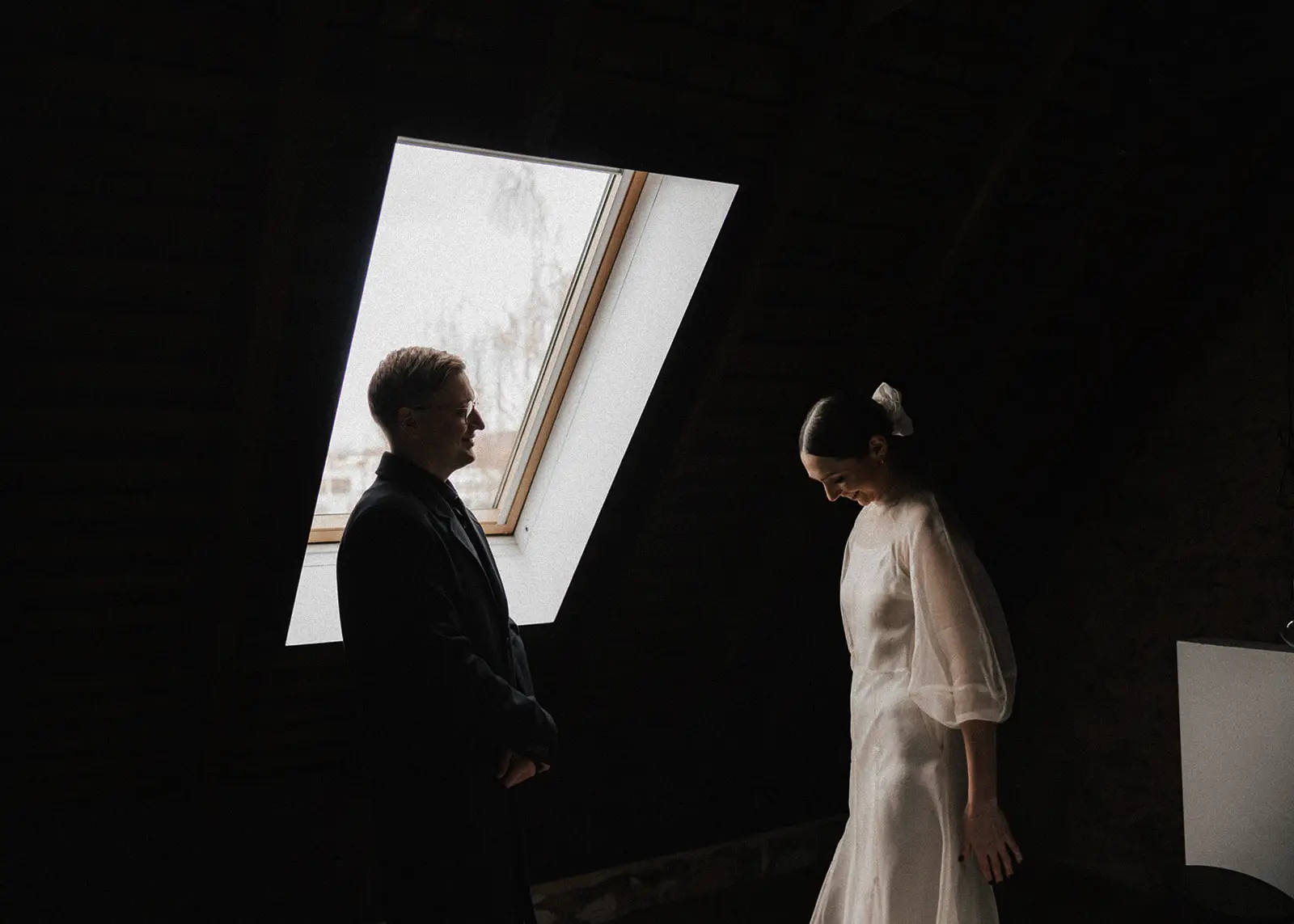 A couple shares an intimate moment inside a historic attic space in Hobart, Tasmania, as soft natural light pours in from a skylight. The bride's delicate gown and groom's dark suit contrast beautifully in this quiet, romantic scene.