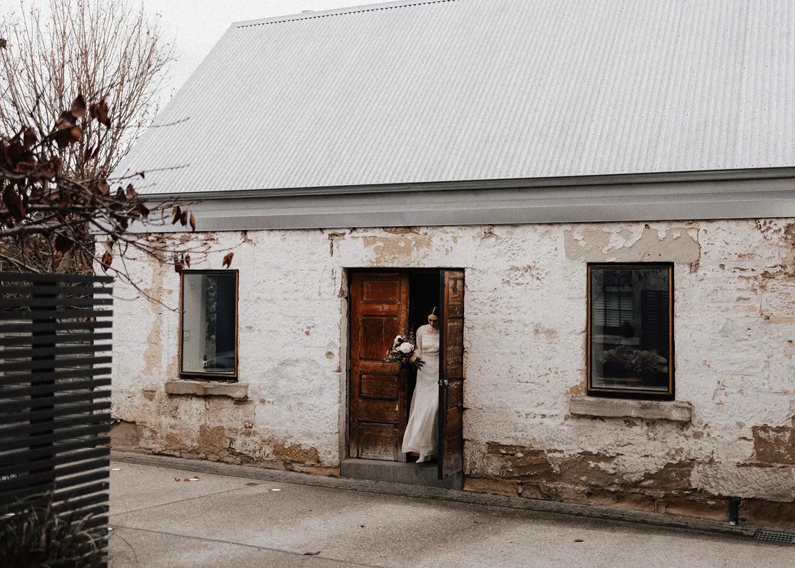 A bride in a flowing wedding dress steps through the rustic wooden doorway of an old stone building in Hobart, Tasmania, carrying a bouquet. The textured walls and historic charm set the scene for a timeless elopement.