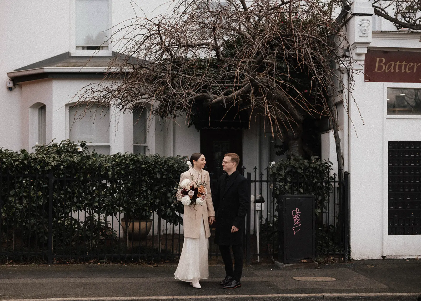 A couple walks together on a Battery Point, Hobart, street in Tasmania, surrounded by heritage buildings and greenery. The bride holds a bouquet of flowers, and both are elegantly dressed, making their way to their intimate elopement.