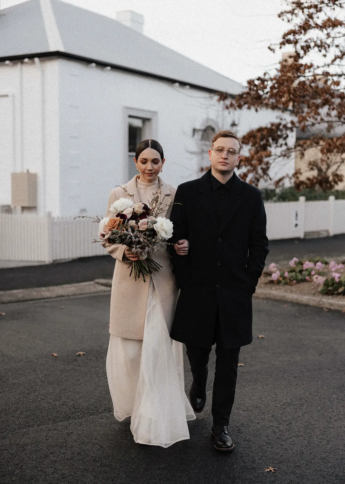 A newlywed couple walks through the charming streets of Battery Point, Hobart, Tasmania, with a soft winter light enhancing their elegant elopement attire. The bride carries a bouquet, and both wear neutral-toned coats over their outfits.