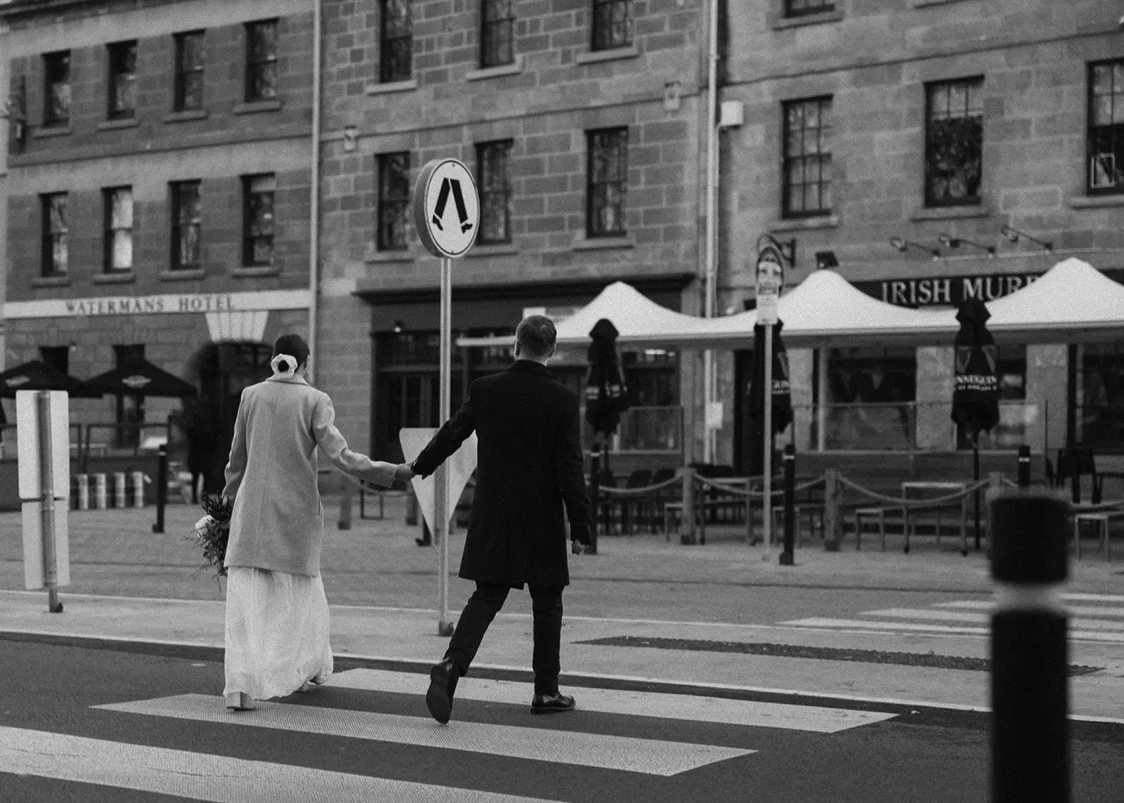 A black and white photograph captures a newlywed couple crossing Salamanca Place in the heart of Hobart, Tasmania, with historic buildings and local pubs in the background, encapsulating the charm of an intimate city elopement.