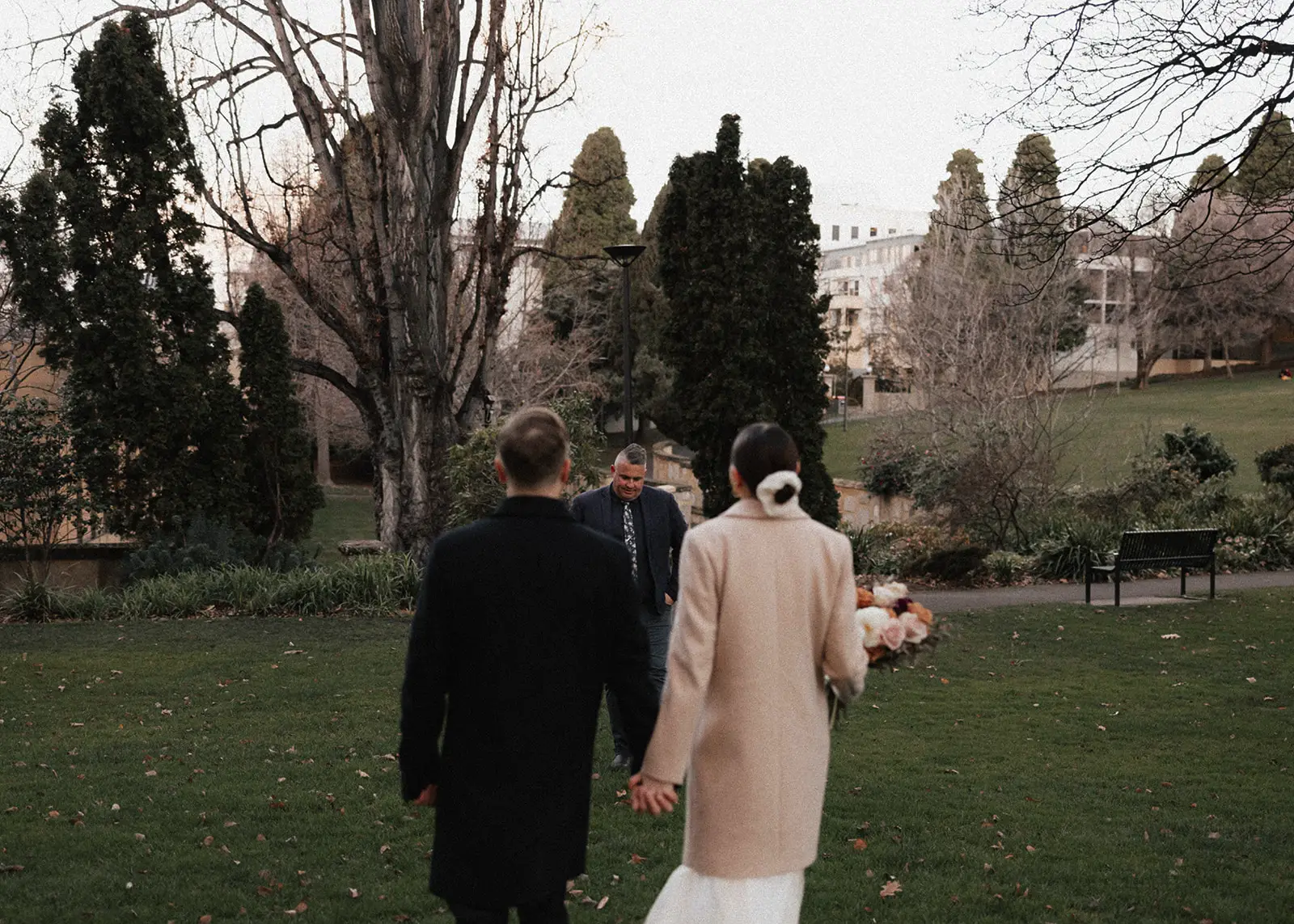 A couple walks hand in hand through a lush green park in Hobart, Tasmania, approaching their wedding celebrant, Josh Withers, for their intimate elopement ceremony. The moment is natural and filled with anticipation.